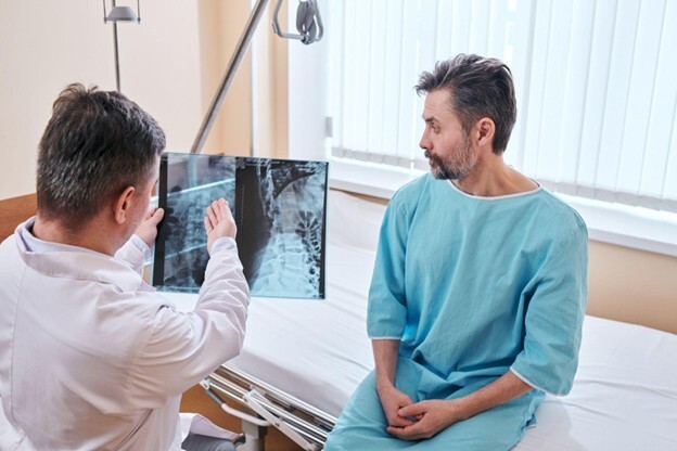Doctor reviewing spinal X-ray with male patient in clinical setting, discussing scoliosis or spinal alignment while patient listens attentively in a medical gown.